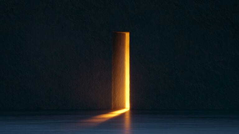 A single narrow doorway, ajar, in a plain dark wall; warm light from the room beyond spills across the floor.